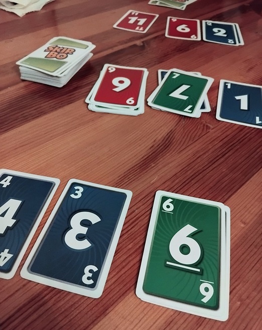 Kuvassa puinen pöytä, jonka päällä osa Skibo-pelikorteista on kuvapuoli ylöspäin, koska peli on kesken. Pelikorteissa on numeroita.