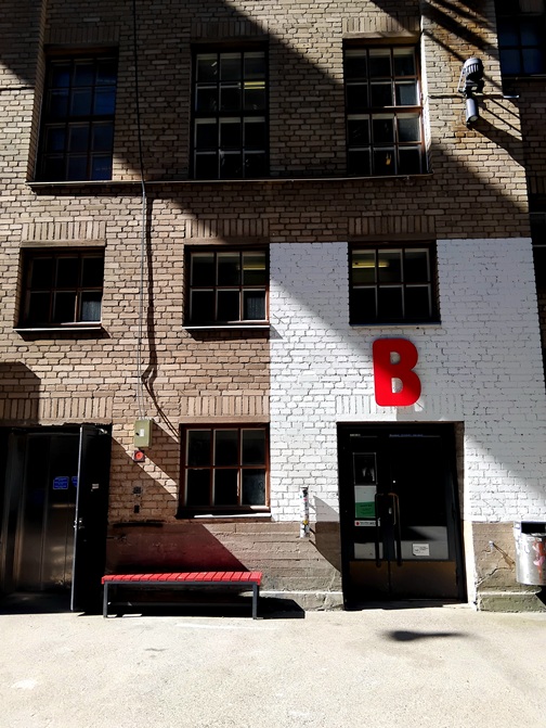 Kaapelitehtaan julkisivu, johon tulee voimakkaita varjoja. B-rapun ympäristö on valkoinen ja siinä on iso punainen B-kirjain