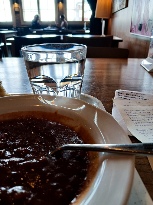 Etualalla keittolautanen, keskiosassa vesilasi, taustalla näkyy ravintolan siluetti ja isot ikkunat