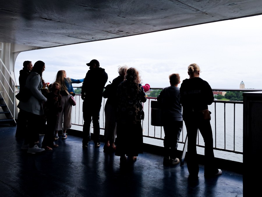 Kuvassa ihmisten siluetteja, heidän katsoessaan laivasta rannikkoa juuri lähdön jälkeen