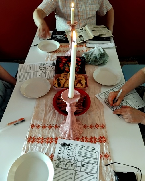 Kuvassa pöytä, jonka äärellä on neljä ihmistä. Pöydällä on peliin liittyviä papereita, syötävää ja 2 kynttilää