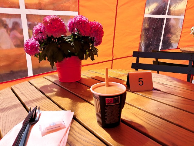 Oranssin teltan sisällä, pöydälle asetettuna kukkia, kahvikuppi ja numerokyltti.