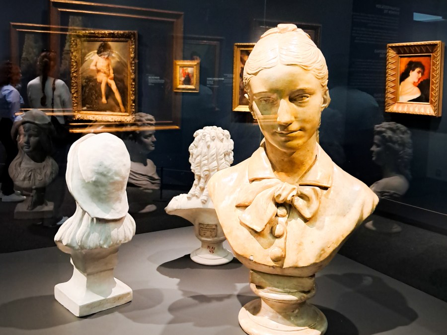 Atemeumin naisten tekemiä potrettipatsaita ja vitriinin heijastuksia. Taustalla museon tauluja ja ihmisiä.