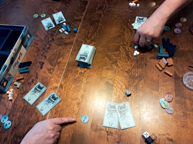 Pöydällä pelikortteja, noppia, pelirahoja sekä pakkauspalikoita, sekä kaksi kättä, jotka järjestelevät pelin aloittamista.