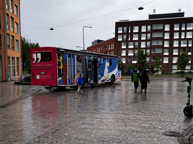 Sateinen kaupunkimiljöö, jossa kirjastoauto ja ihmisiä.